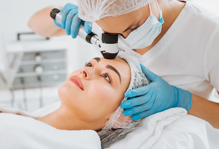 Stock photograph of models: a masked clinician in a hair cover checks a reclining woman's forehead with a dermatoscope