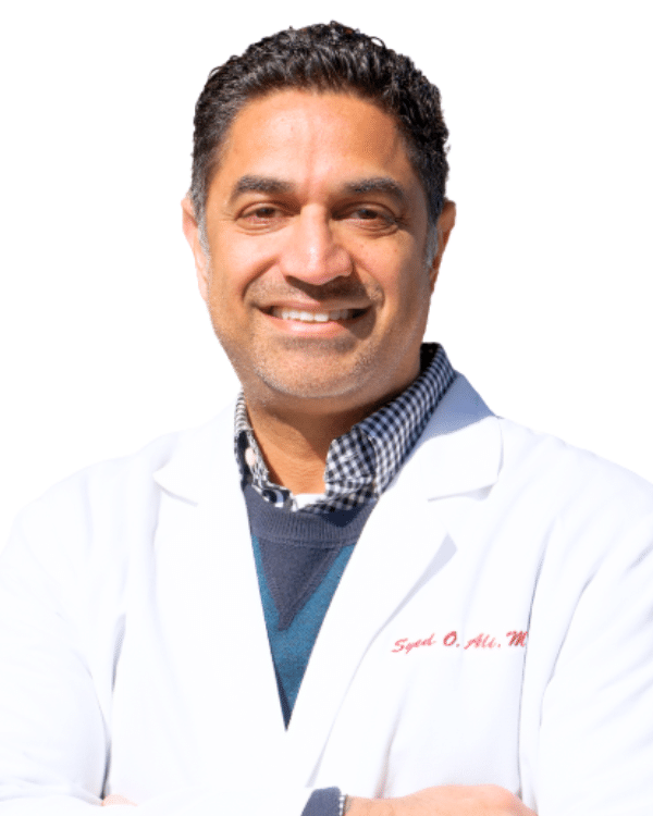 Dr. Syed O. Ali, smiling, in a white coat embroidered with his name over a checked shirt and blue sweater
